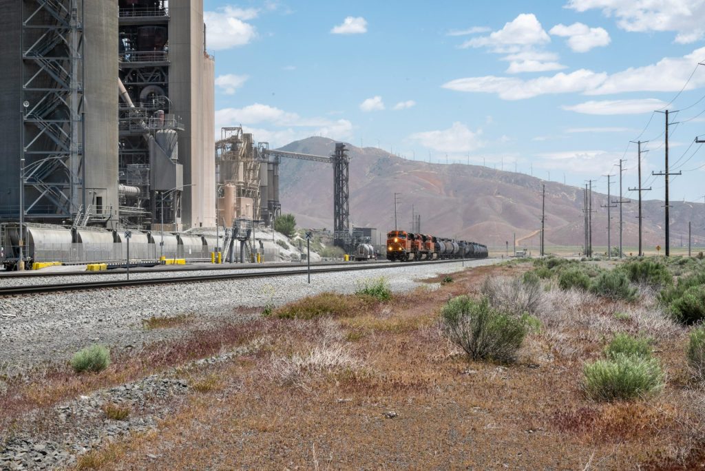 Freight train passing a large industrial facility in Monolith, CA, with mountains in the background.